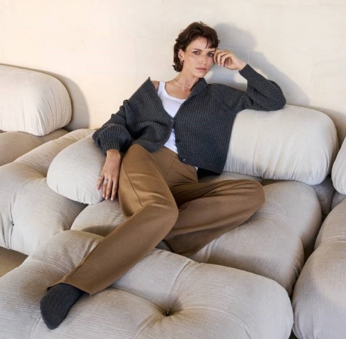 žena sedící na gauči v bílém tílku, šedém svetru a hnědých kalhotách