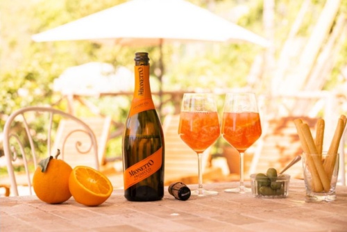 Osobní vinotéka - na stole otevřená láhev alkoholu, dvě sklenice s nápoji, tyčinky ve sklenici a pomeranče