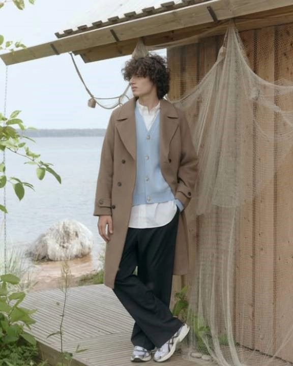 muž v hnědém kabátu, košili a černých kalhotách stojící na verandě u domu