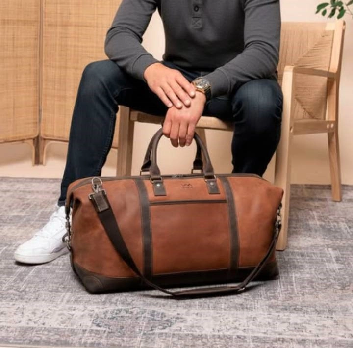 muž v černém oblečení sedící na židli a v ruce držící hnědou cestovní tašku